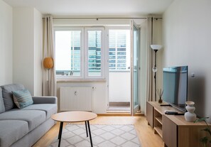 Ein modernes Wohnzimmer mit einem grauen Sofa, einem kleinen runden Tisch, einem Fernseher und Blick auf eine Stadtansicht durch das Fenster.