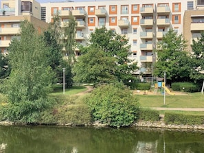 Ein Wohngebäude mit mehreren Balkonen, ein Park mit Spielplatz und ein Gewässer im Vordergrund.