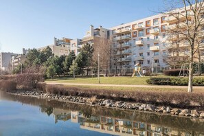 A residential building with balconies, a park with a pond, and a clear sky. - Wyględowska 2-Bedroom Apartment (Warszawa)
