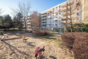 A playground with sand and play equipment, surrounded by trees and a multi-story building with balconies. - Wyględowska 2-Bedroom Apartment (Warszawa)