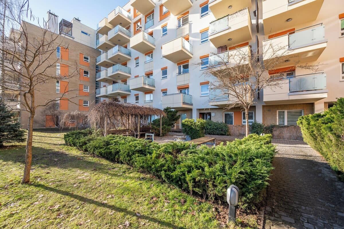 Un immeuble résidentiel à plusieurs étages, doté de balcons, d’un espace jardin agrémenté d’arbustes et d’un chemin bordé de briques.