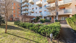 Um edifício residencial com vários andares, varandas, área de jardim com arbustos e um caminho de tijolos.