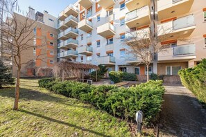 Een meerlaags woongebouw met balkons, een tuin met struiken en een met baksteen bestraat pad.