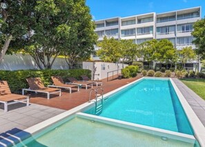 A swimming pool with lounge chairs and a modern building in the background. - Coastal Escape | Heart of Nelson Bay | Family-Friendly Poolside Paradise (Nelson Bay)