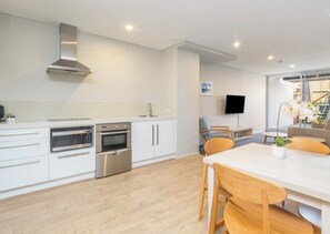 A modern kitchen with white cabinetry, stainless steel appliances, and a wooden dining table with chairs. - Coastal Escape | Heart of Nelson Bay | Family-Friendly Poolside Paradise (Nelson Bay)