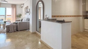 A modern living room with a sofa and a coffee table, a kitchen with a countertop and a mirror, and a hallway with a plant. - Pink La Perla Apartment (Zenia, La)