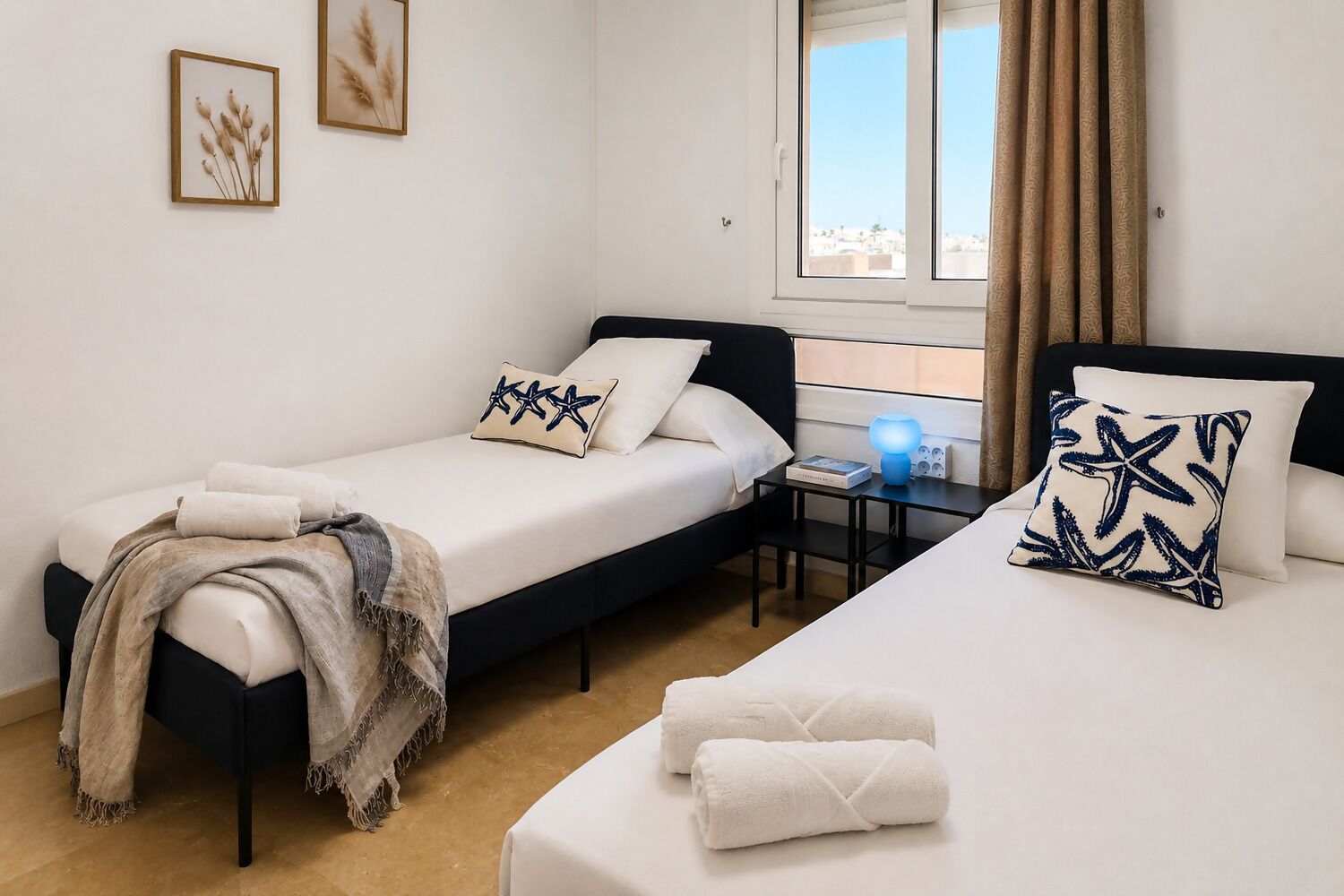 A hotel room with two single beds, a desk with a lamp, and a window with curtains.