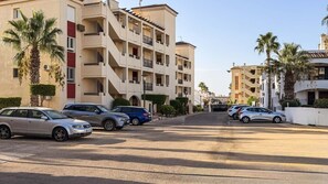 Un edificio residencial de varios pisos con balcones, coches aparcados y palmeras.