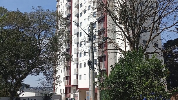 Un immeuble résidentiel de grande hauteur doté de nombreux balcons, un espace de stationnement avec des voitures et des motos, et une rue bordée d’arbres.
