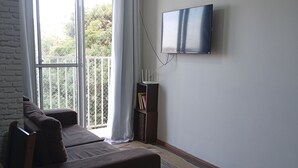 Un salon comprenant un canapé, un téléviseur à écran plat fixé au mur, une fenêtre avec des rideaux et un balcon doté d’une balustrade.