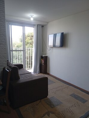 A living room with a sofa, a flat-screen TV mounted on the wall, a window with curtains, and a balcony with a railing. - Cozy apartment with complete security in the Serra Gaúcha region, within the UniverUCS campus. (Caxias do Sul)