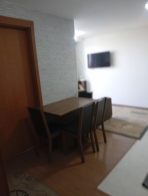 A room with a wooden dining table, chairs, a white brick wall, and a mounted television. - Cozy apartment with complete security in the Serra Gaúcha region, within the UniverUCS campus. (Caxias do Sul)