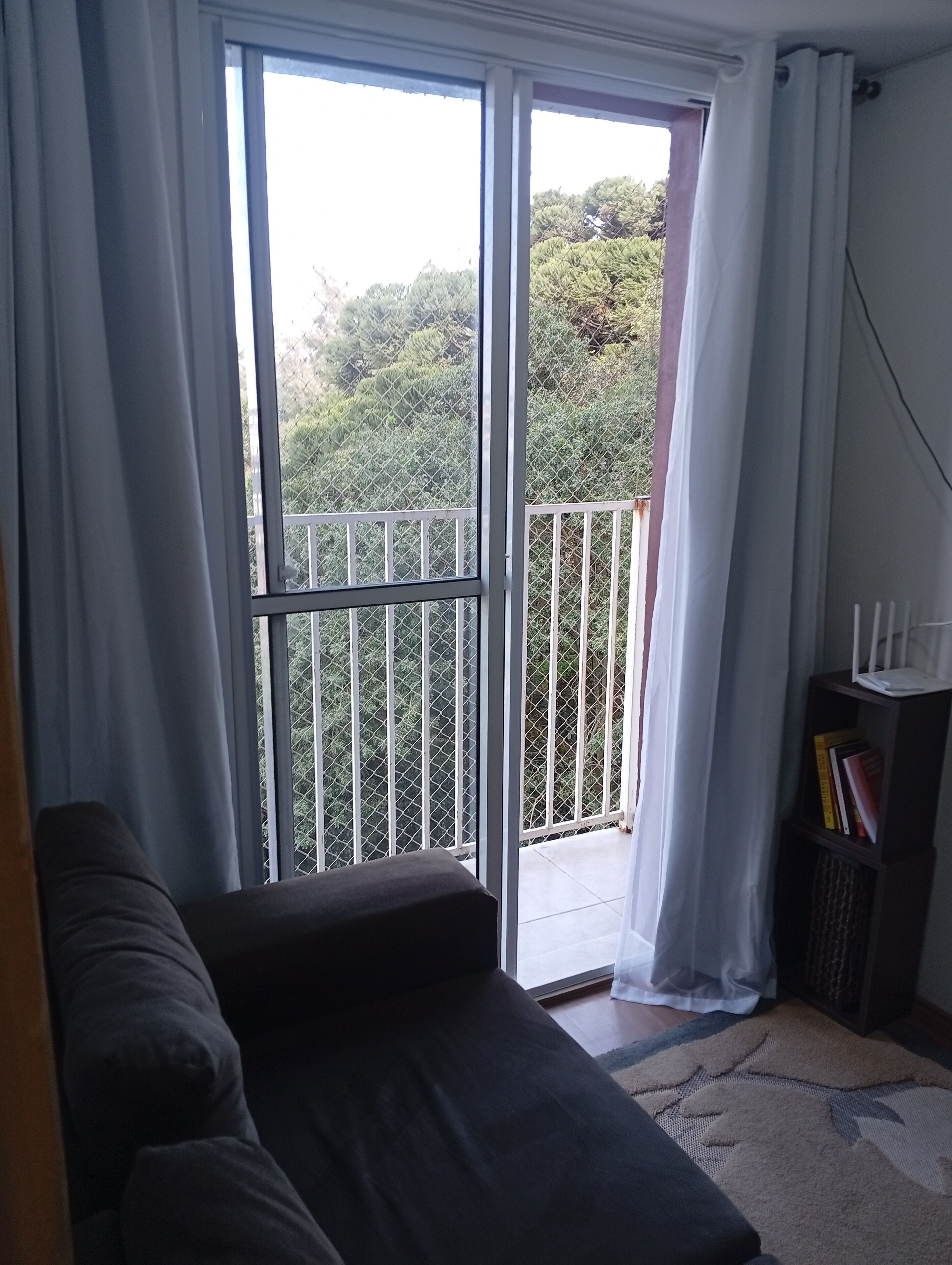 Quarto com um sofá escuro, uma pequena estante e vista para uma varanda com guarda-corrimão de metal através de uma porta de correr de vidro.