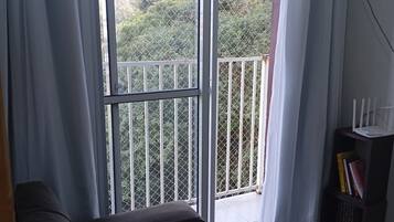 Quarto com um sofá escuro, uma pequena estante e vista para uma varanda com guarda-corrimão de metal através de uma porta de correr de vidro.