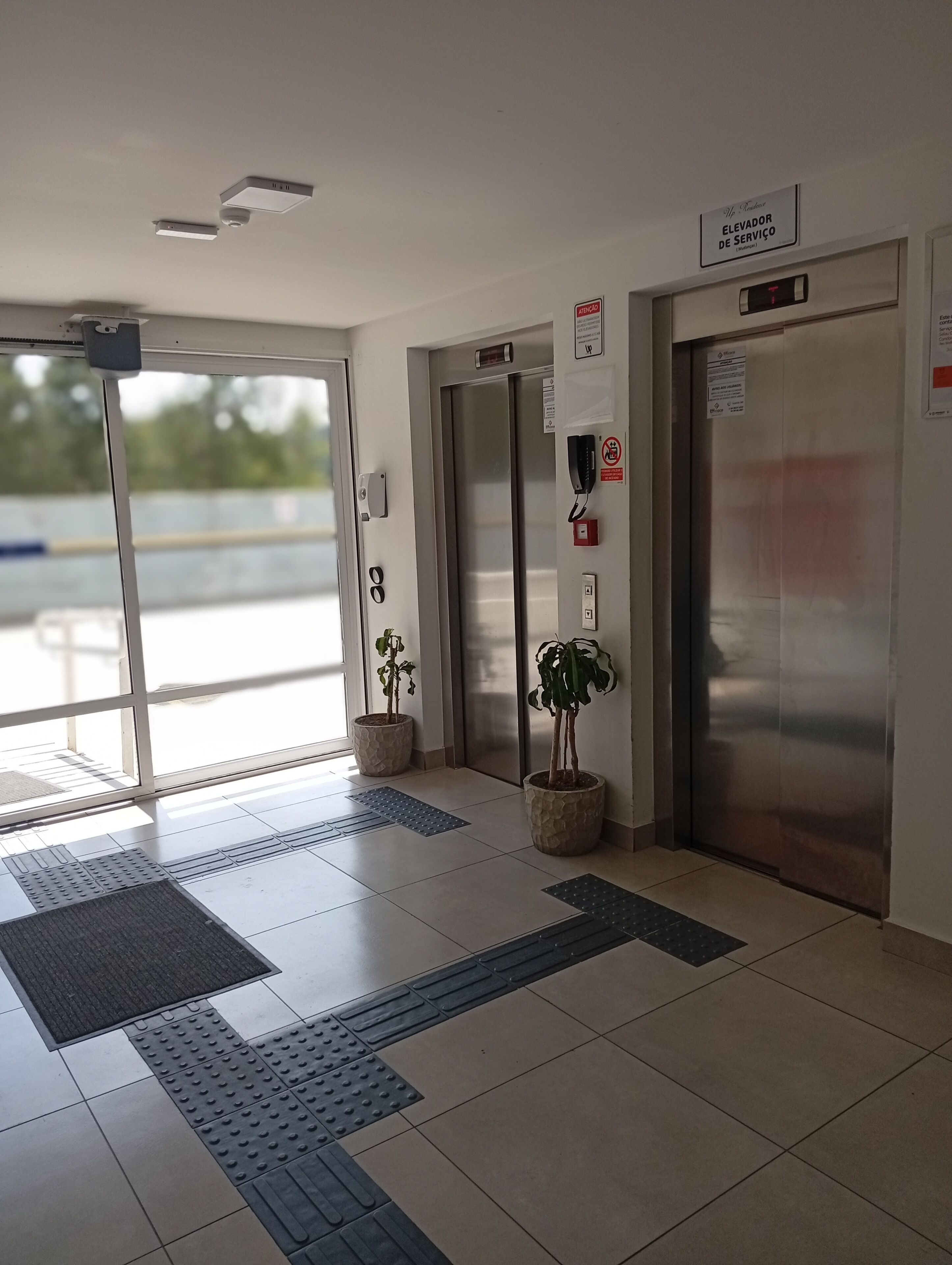 An elevator area with two elevators, a fire alarm, a telephone, and tactile paving on the floor.