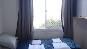Un lit simple avec un matelas bleu et du linge de lit blanc, une fenêtre avec des rideaux et une étagère fixée au mur.