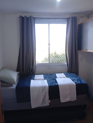 A single bed with a blue mattress and white linens, a window with curtains, and a wall-mounted shelf. - Cozy apartment with complete security in the Serra Gaúcha region, within the UniverUCS campus. (Caxias do Sul)