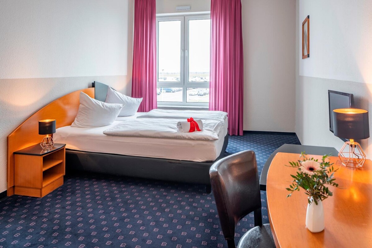 Habitación de hotel con cama, escritorio con silla, una mesita con un florero y ventana con cortinas.