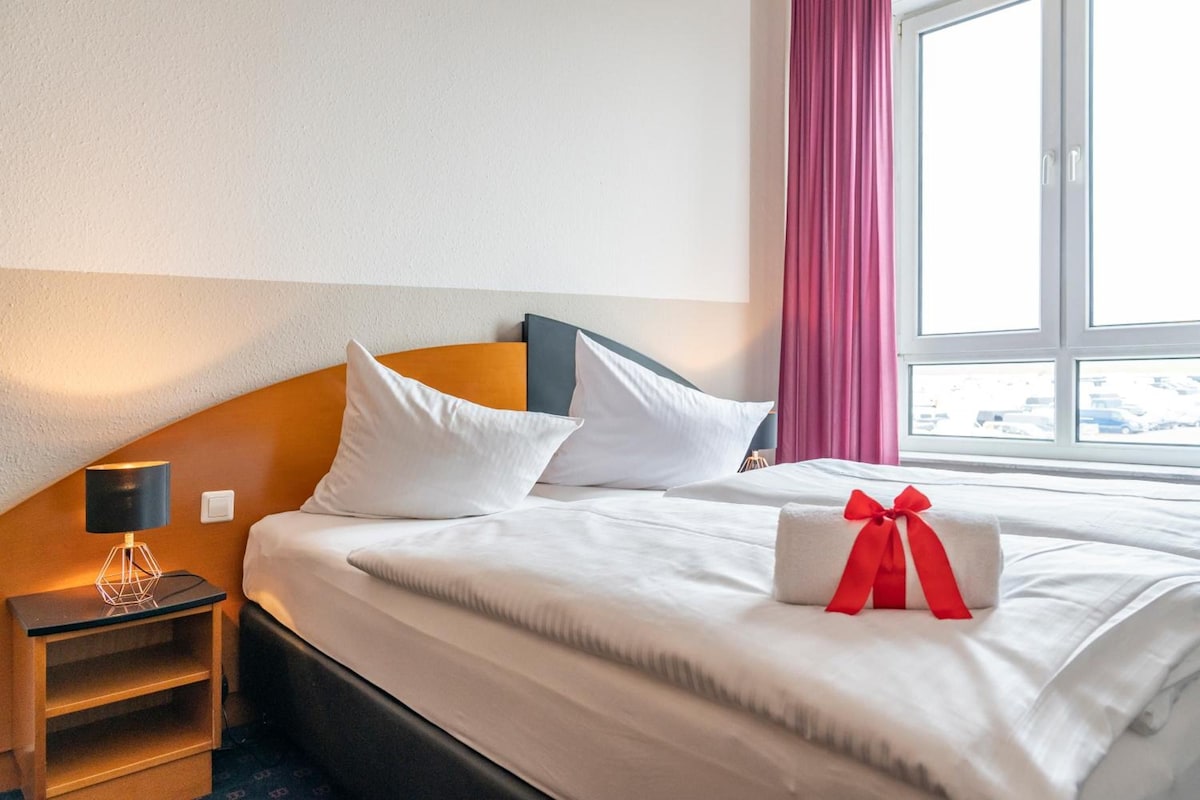 Habitación de hotel con cama, almohadas, mesita de noche, lámpara y ventana con cortinas.