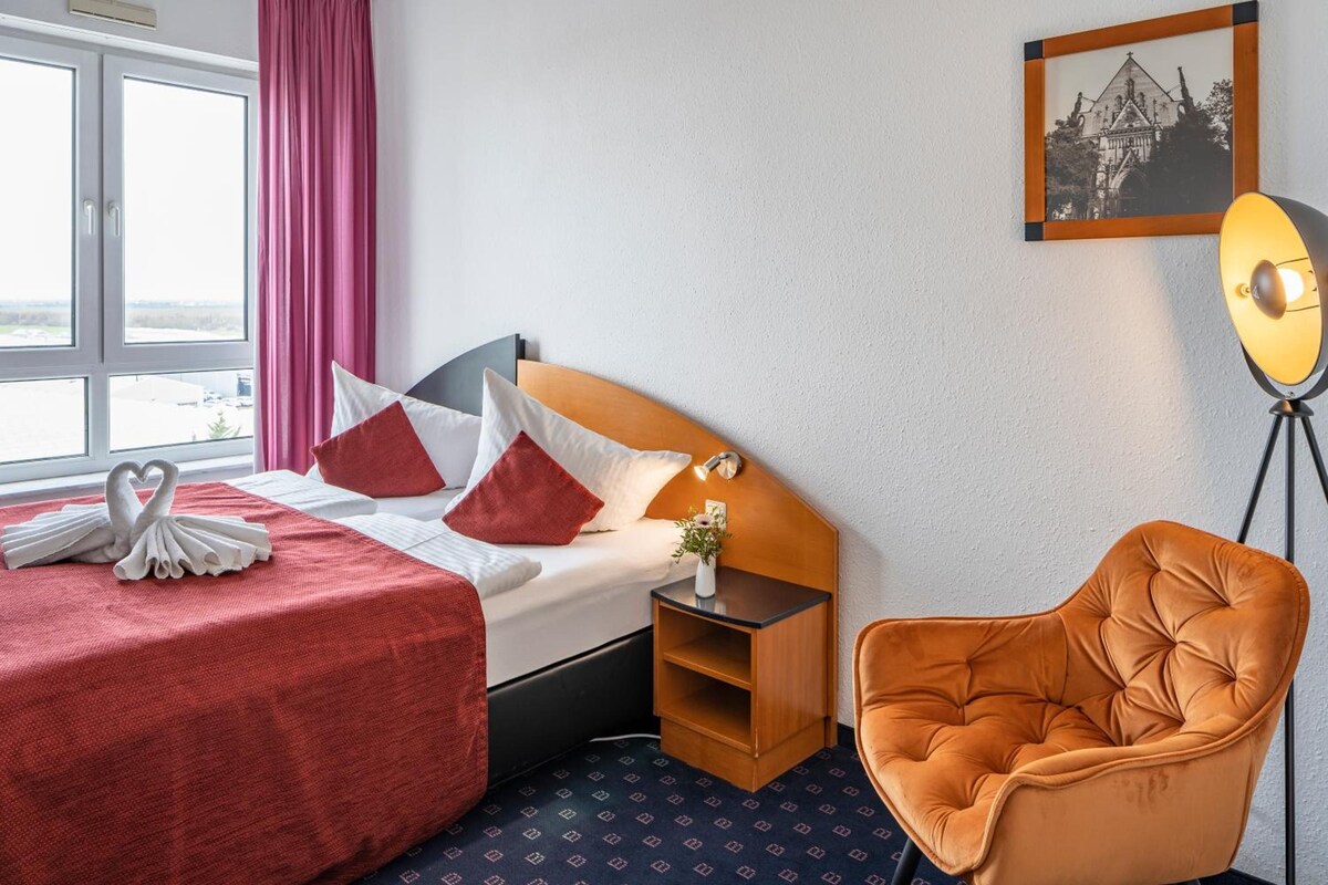Una habitación de hotel con cama, mesita de noche, silla, lámpara y un cuadro en la pared.