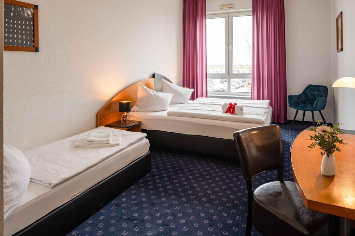 Habitación de hotel con dos camas individuales, una mesita con un florero y una silla.