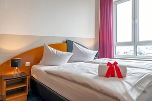 Een hotelkamer met een bed, kussens, een nachtkastje, een lamp en een raam met gordijnen.