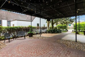 A covered outdoor area with a brick pathway, benches, potted plants, and a building in the background.