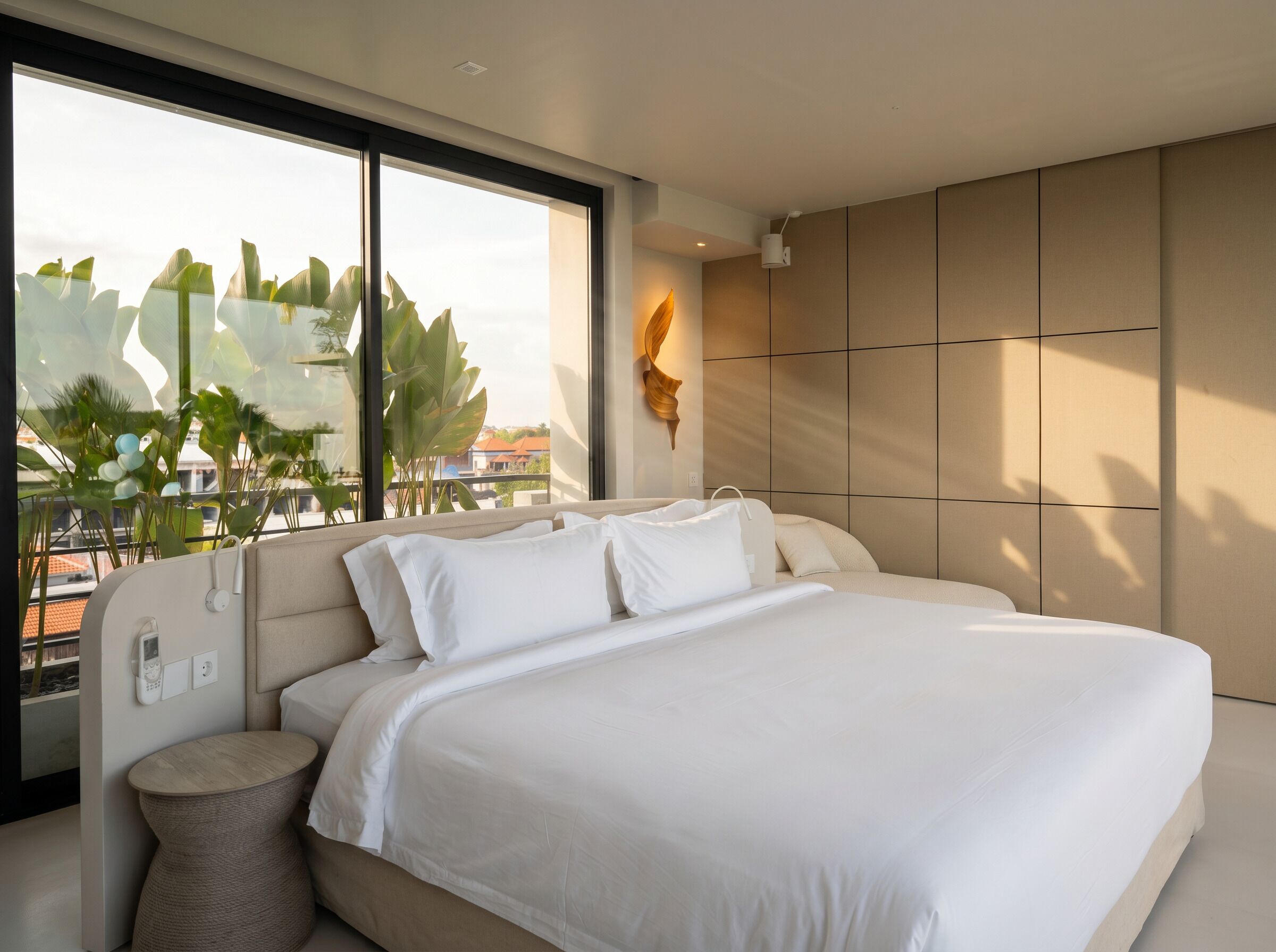 Une chambre moderne avec un grand lit, une table de chevet, une lampe fixée au mur et une vue sur l’extérieur.