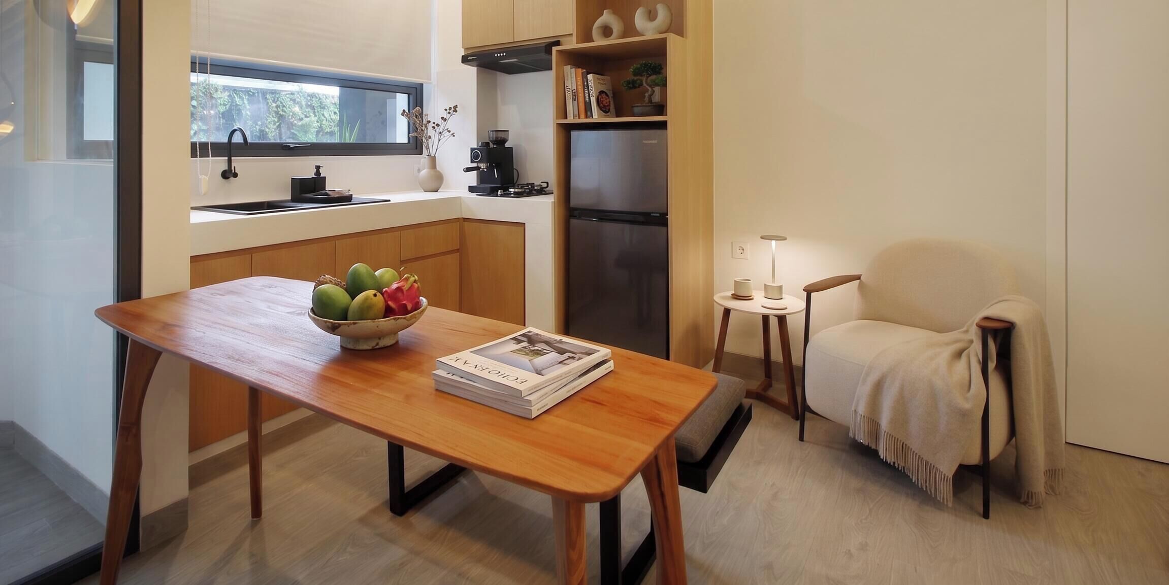A compact kitchen with a wooden table, a bowl of fruit, a small side table with a lamp, and a chair with a blanket.