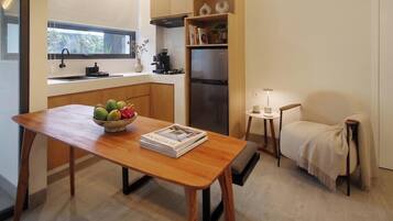 A compact kitchen with a wooden table, a bowl of fruit, a small side table with a lamp, and a chair with a blanket.