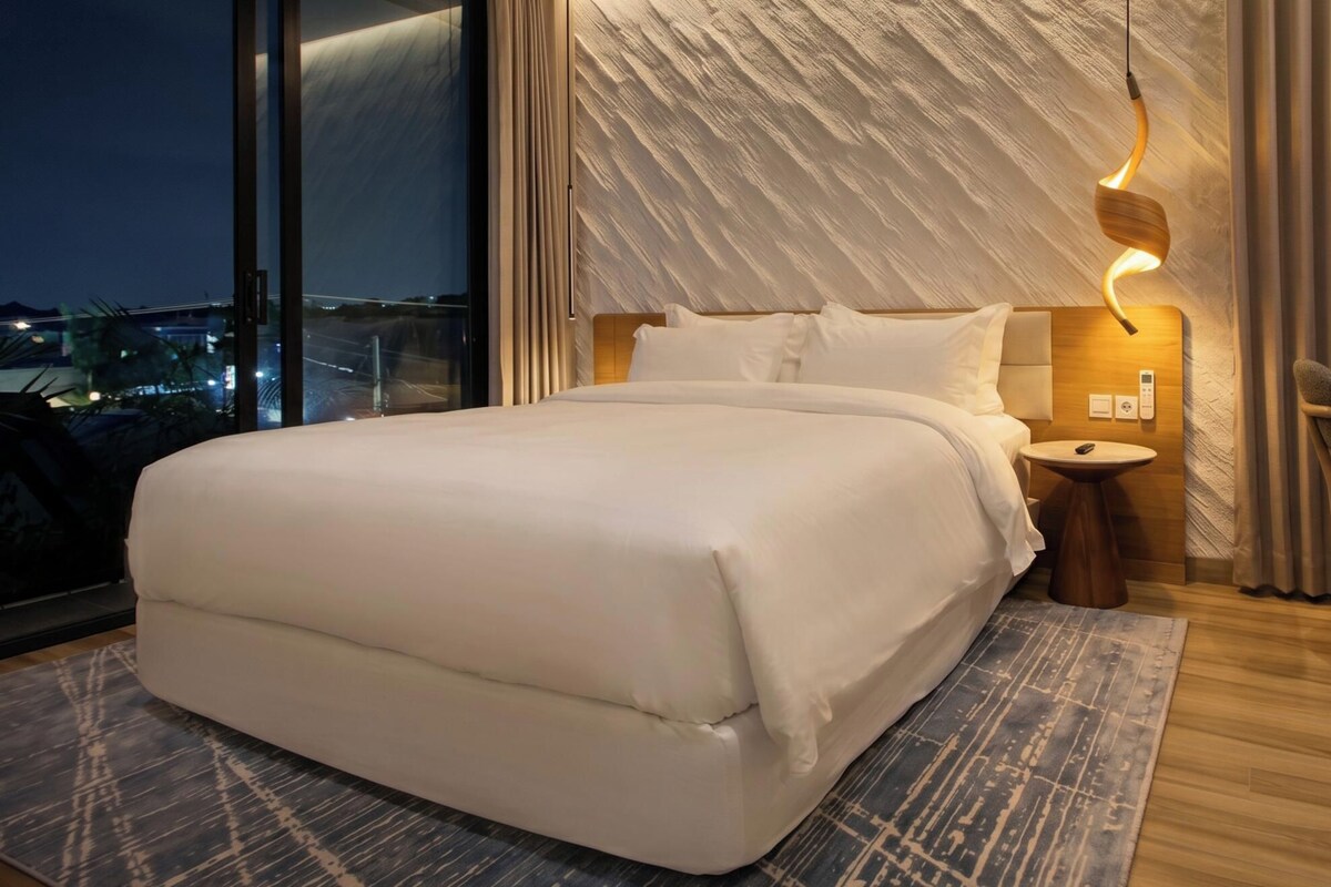 Habitación de hotel moderna con una cama grande, una mesita de noche, una pequeña mesa redonda, un florero y vistas nocturnas de la ciudad.
