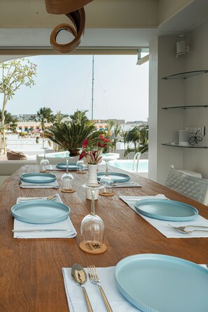 Mesa de comedor dispuesta con platos azules, cubiertos y copas de vino, con vistas a una piscina y palmeras.