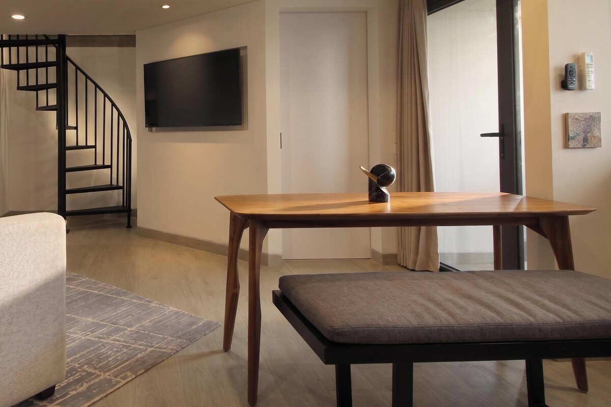 Una sala moderna con una mesa de madera, un banco, una escalera y un televisor de pantalla plana montado en la pared.