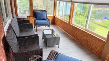 A sunlit room with wicker furniture, a blue cushioned chair, and a view of a lake through large windows.
