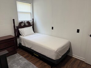 A small, single-bed room with a wooden headboard, a nightstand, a dresser, and a window with blinds.