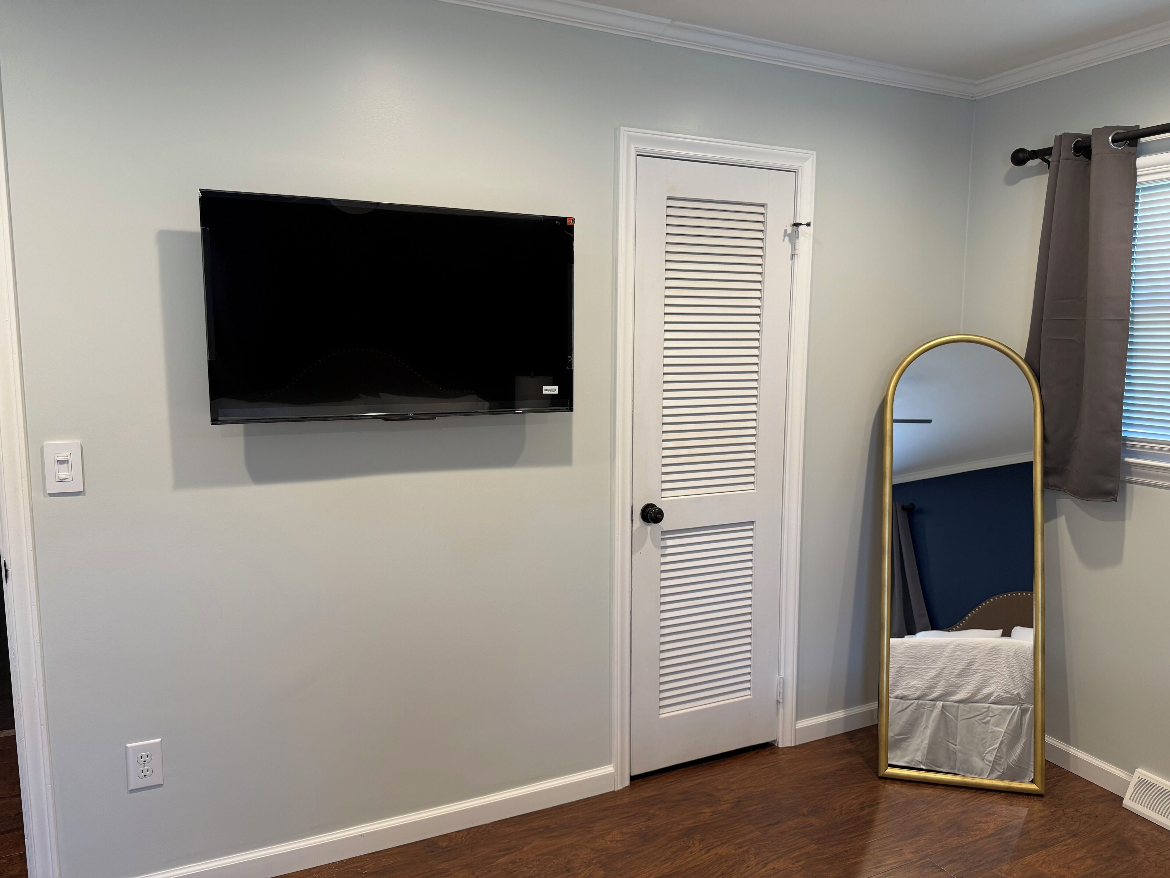 Een kamer met een witte deur, een wandgemonteerde flatscreen televisie, een spiegel in geheel lengte met een gouden lijst en een stopcontact aan de muur.