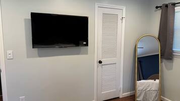Een kamer met een witte deur, een wandgemonteerde flatscreen televisie, een spiegel in geheel lengte met een gouden lijst en een stopcontact aan de muur.