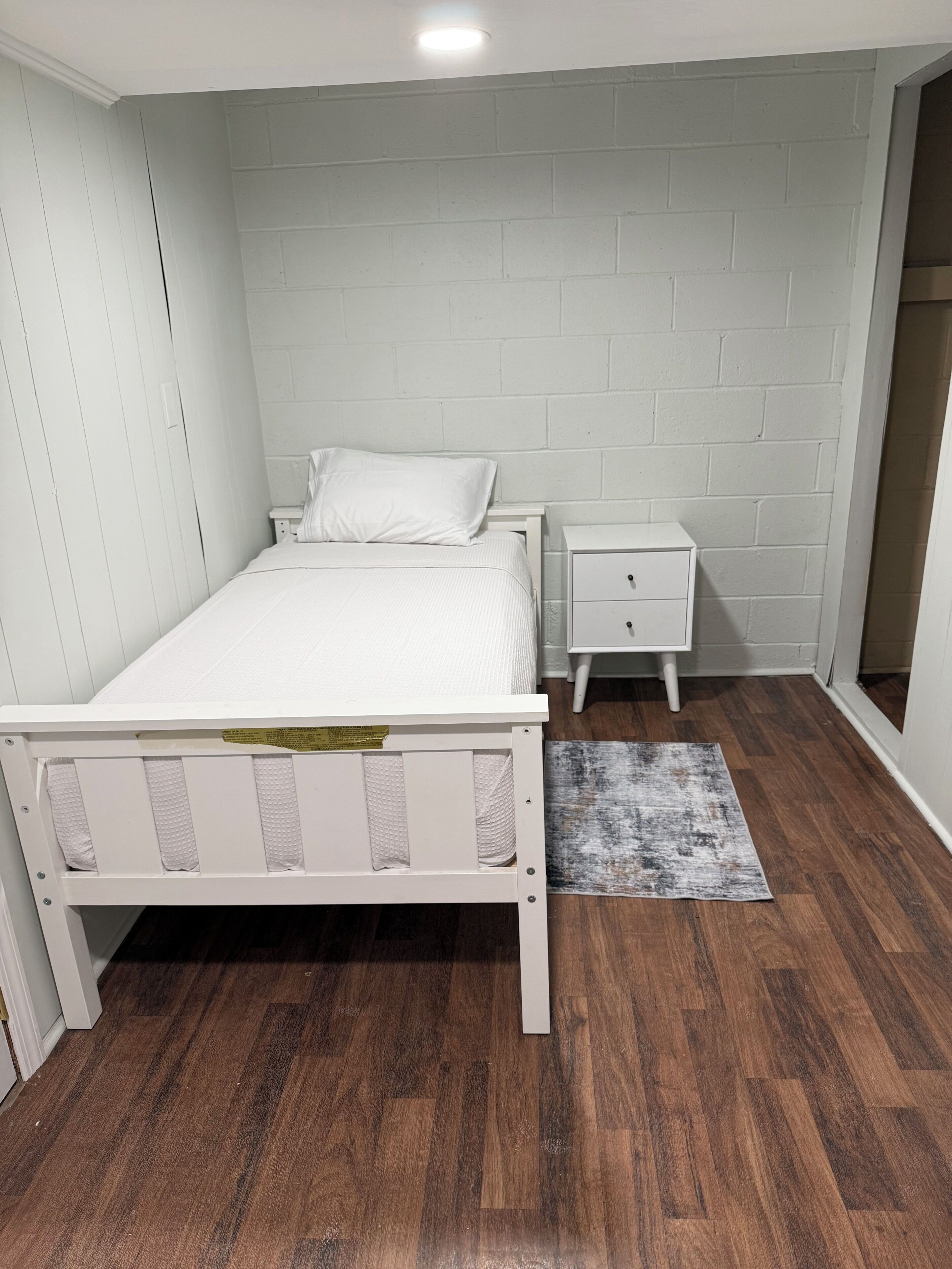 Een slaapkamer met een wit bed, een nachtkastje, een klein lade, een houten vloer en een witte baksteenmuur.