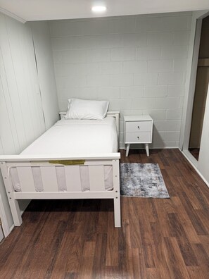 A bedroom with a white bed, a nightstand, a small drawer, a wooden floor, and a white brick wall.
