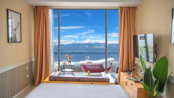 Habitación de hotel con una cama grande, una zona de estar con mesa y sillas, una bañera y vistas al mar y a las montañas.