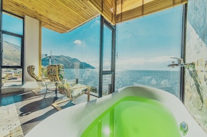 A modern bathroom with a large bathtub, a seating area with chairs, and a view of the sea.