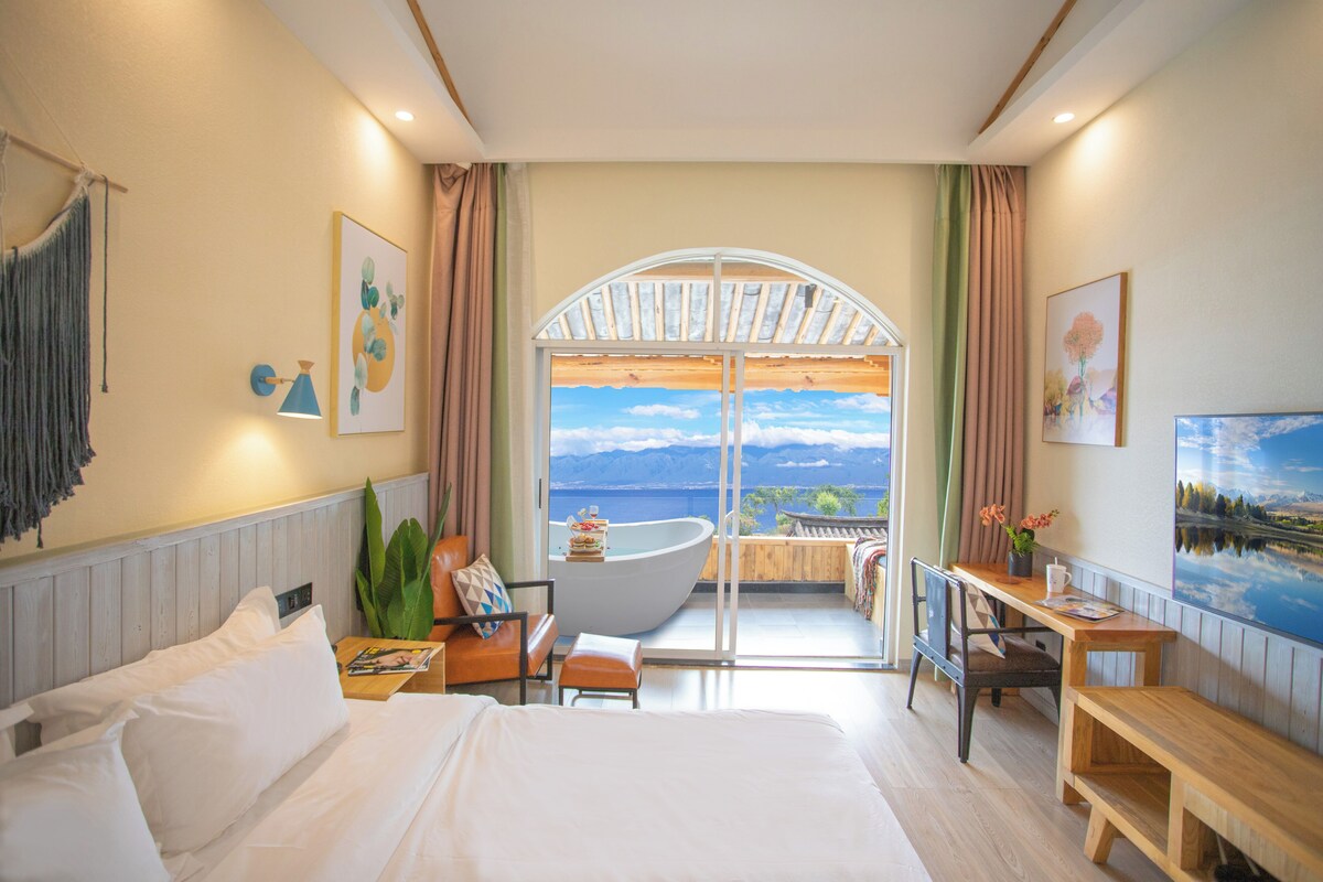 A hotel room with a large bed, a desk, a chair, a balcony with a view of the mountains and water.