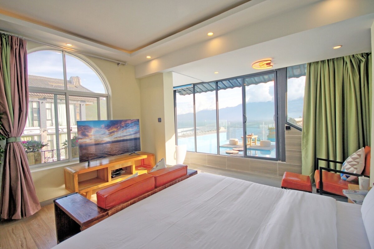 A hotel room with a large bed, a television, a seating area, and a balcony with a pool view.