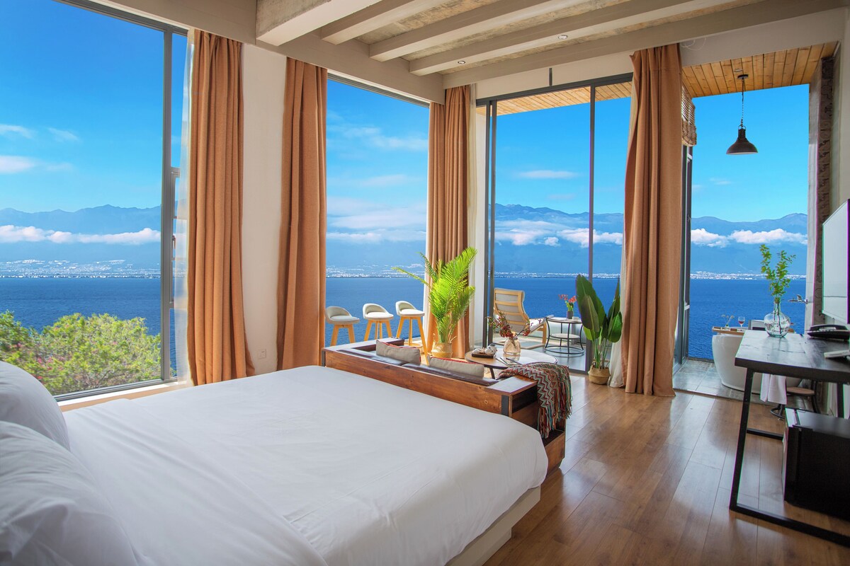 A hotel room with a large bed, a dining area, and a view of the sea.