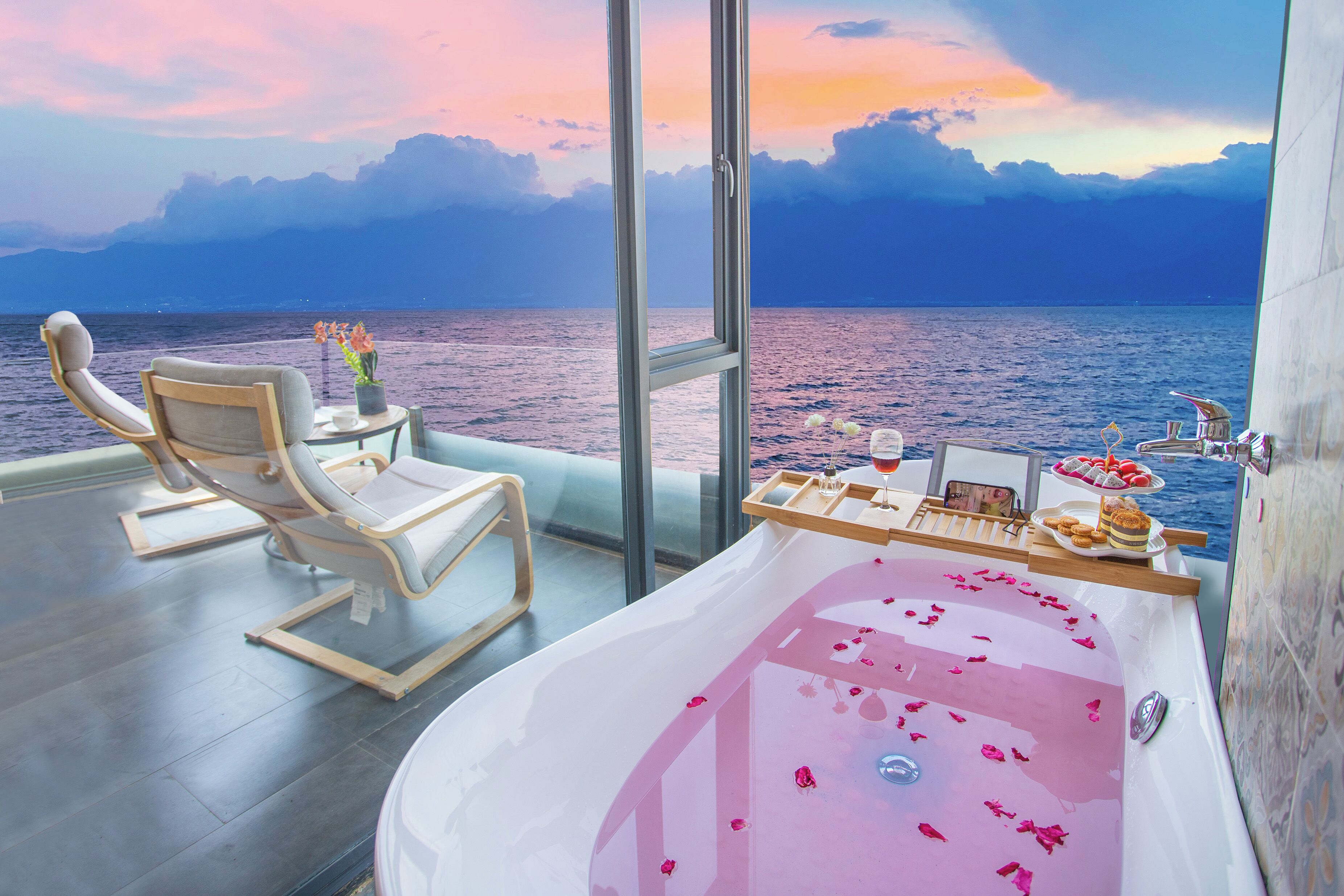 A balcony with a view of the sea, a bathtub with rose petals, and a table with snacks and a drink.