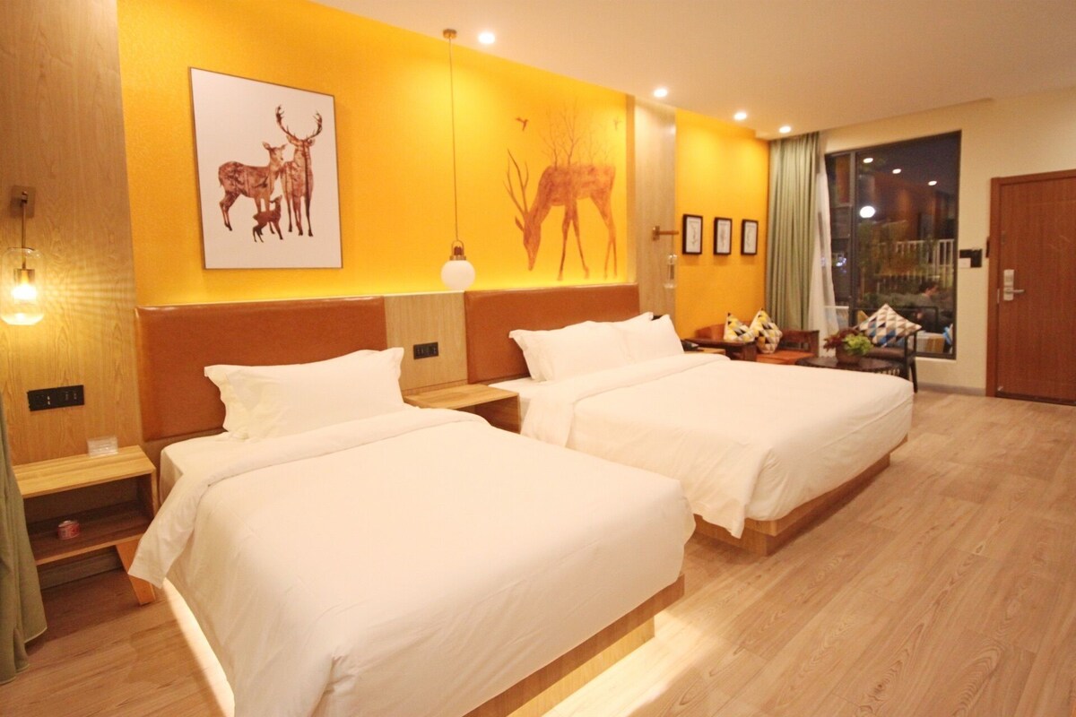 A hotel room with two beds, a nightstand, a lamp, and a window with a view.