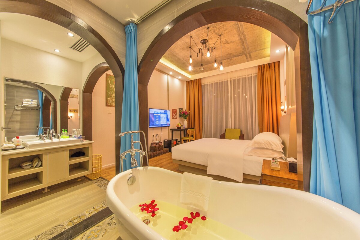 A hotel room with a large bathtub, a bed, a desk with a computer, and a bathroom with a mirror and sink.