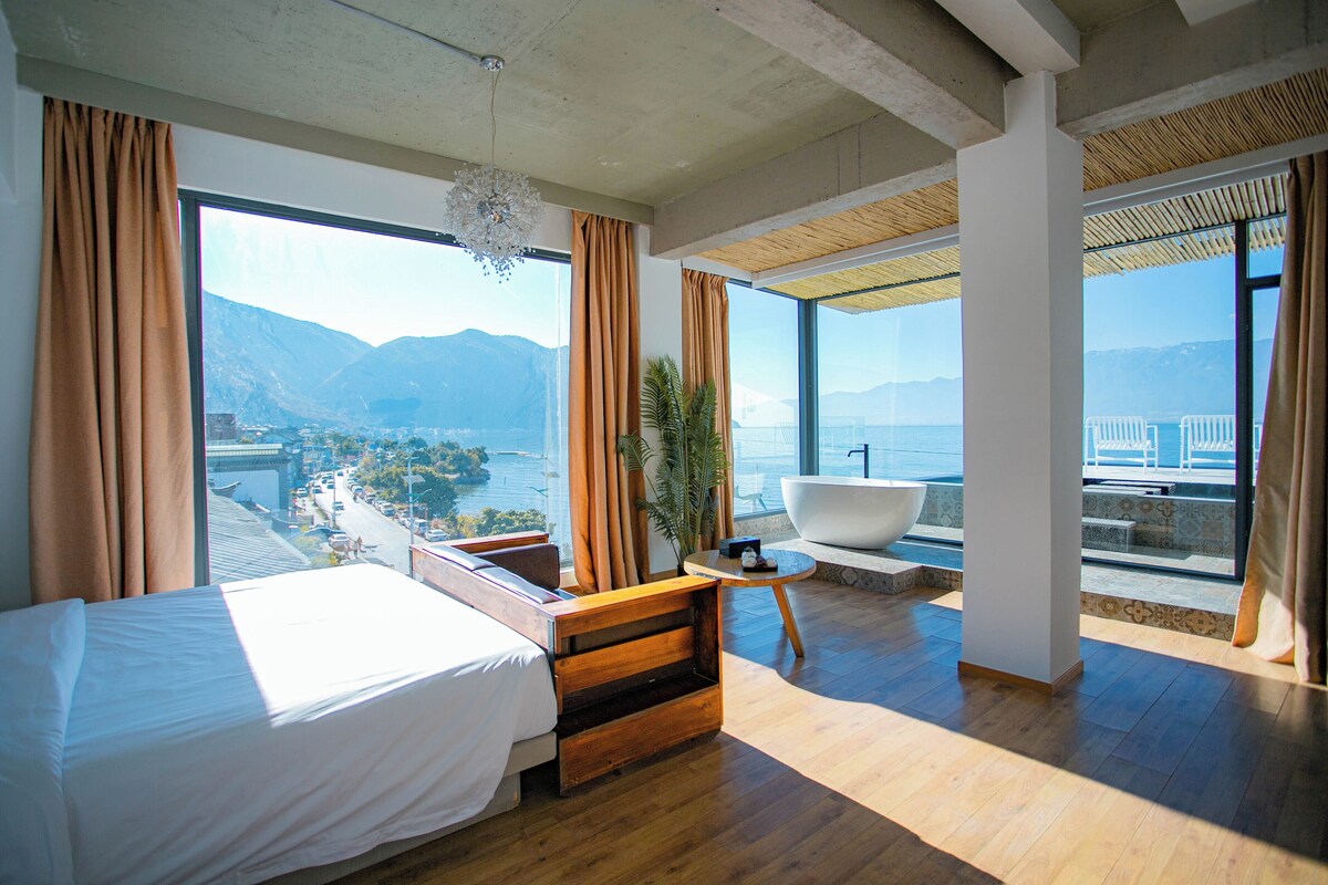 A hotel room with a bed, a seating area with a view of the sea, and a large window overlooking mountains.