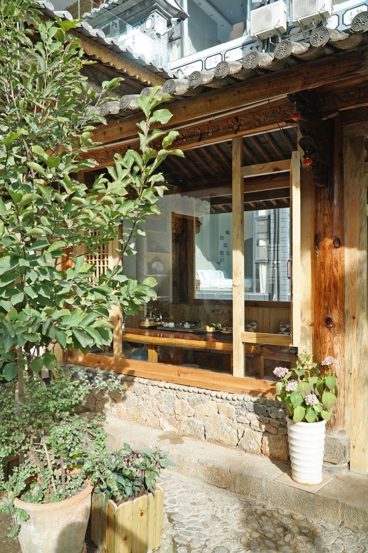 Un edificio tradicional de madera con base de piedra, escalones de madera que conducen a una puerta de madera, y una maceta con flores rosas en el frente.