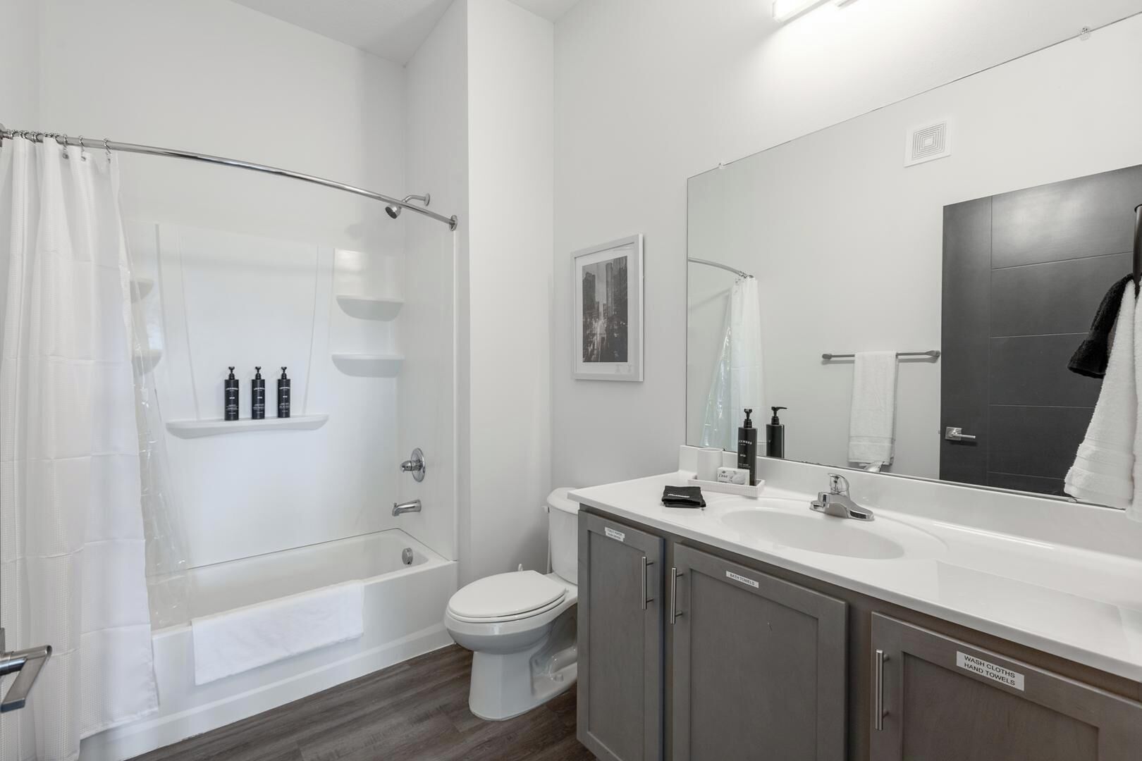 A modern bathroom with a glass shower enclosure, a white toilet, a vanity with a mirror, and a cabinet labeled for wash cloths and hand towels.